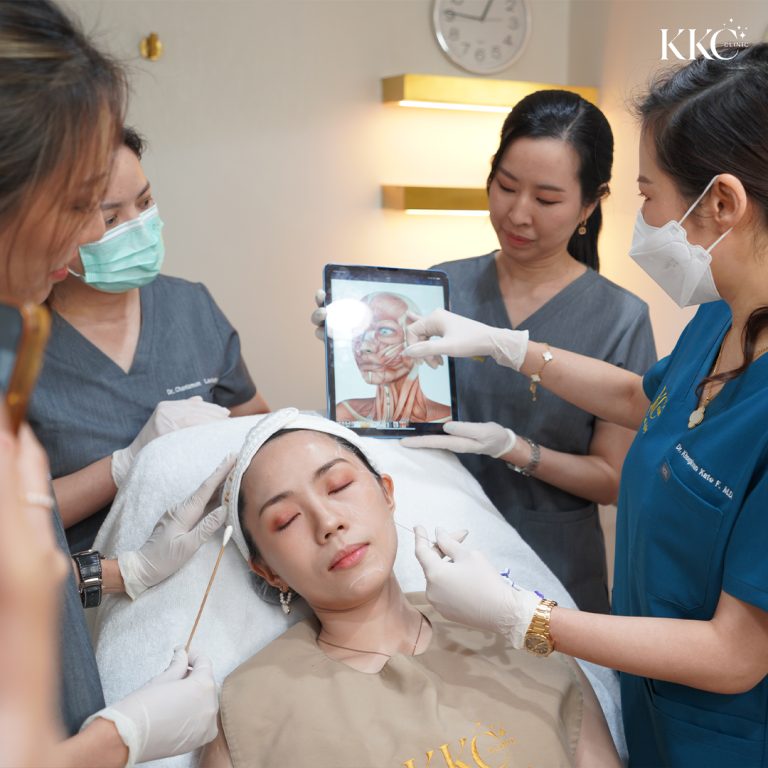 ผู้ป่วยนอนหลับตาระหว่างการทำหัตถการ Asian MD Codes ที่ KKC Clinic โดยมีทีมแพทย์หลายคนล้อมรอบ หนึ่งในนั้นถือ iPad ที่แสดงภาพกายวิภาคของกล้ามเนื้อใบหน้า ขณะที่ทีมแพทย์กำลังพูดคุยเกี่ยวกับการทำหัตถการ และหนึ่งในนั้นกำลังเตรียมฉีดยา