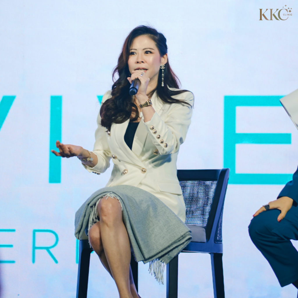 พญ.ของขวัญ ฟูจิตนิรันดร์ กำลังพูดผ่านไมโครโฟนขณะนั่งบนเวทีในงาน Skinvive โดยสวมเสื้อเบลเซอร์สีขาวและกระโปรงสีเทาอ่อน