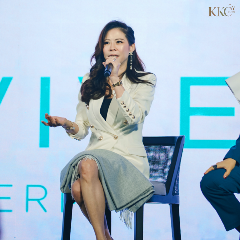 พญ.ของขวัญ ฟูจิตนิรันดร์ กำลังพูดผ่านไมโครโฟนขณะนั่งบนเวทีในงาน Skinvive โดยสวมเสื้อเบลเซอร์สีขาวและกระโปรงสีเทาอ่อน