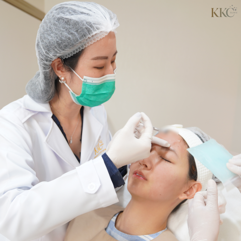 แพทย์ประจำ KKC Clinic สวมหน้ากากสีเขียวและชุดกาวน์สีขาว กำลังฉีดโบท็อกซ์บริเวณหน้าผากผู้ป่วย โดยมีผู้ช่วยถือแผ่นประคบเย็นอยู่ด้านข้าง