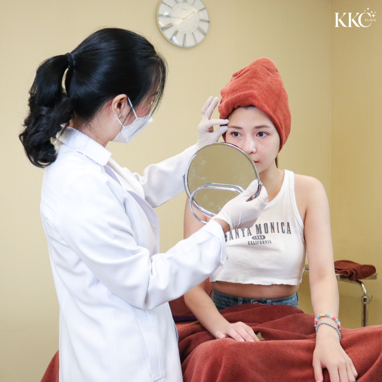 แพทย์ประจำ KKC Clinic สวมชุดกาวน์และถุงมือ กำลังให้คำปรึกษาผู้ป่วยที่มีผ้าขนหนูสีแดงคลุมศีรษะ ผู้ป่วยถือกระจกขณะถูกตรวจบริเวณหน้าผาก