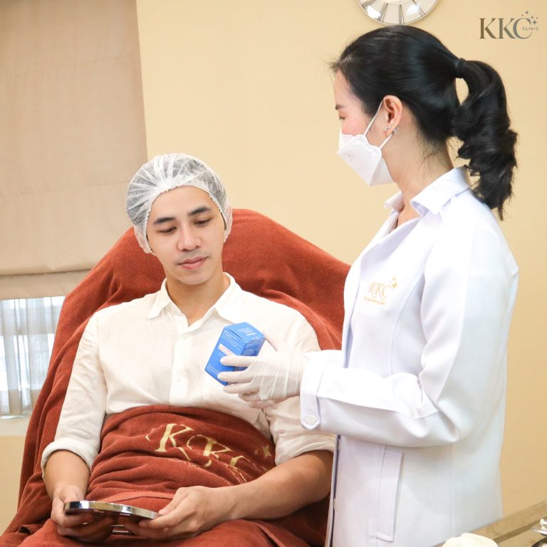 ผู้ชายกำลังนั่งบนเก้าอี้รักษา มีผ้าห่ม KKC คลุมตัว ถือกระจกในมือ ขณะที่ผู้เชี่ยวชาญในชุดคลุมสีขาวและสวมถุงมือกำลังแสดงกล่อง Xeomin ให้ดู