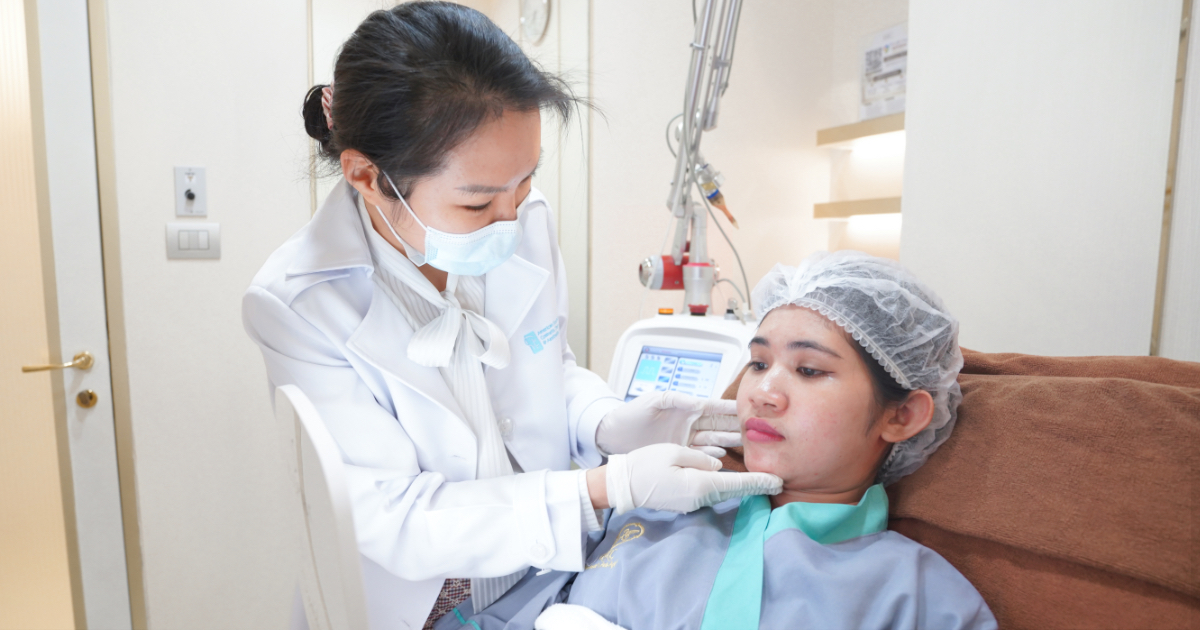 A doctor wearing a white coat, face mask, and gloves examining a female patient’s chin during a skincare consultation. The patient is wearing a disposable hair cover and a clinic gown while reclining on a treatment chair.