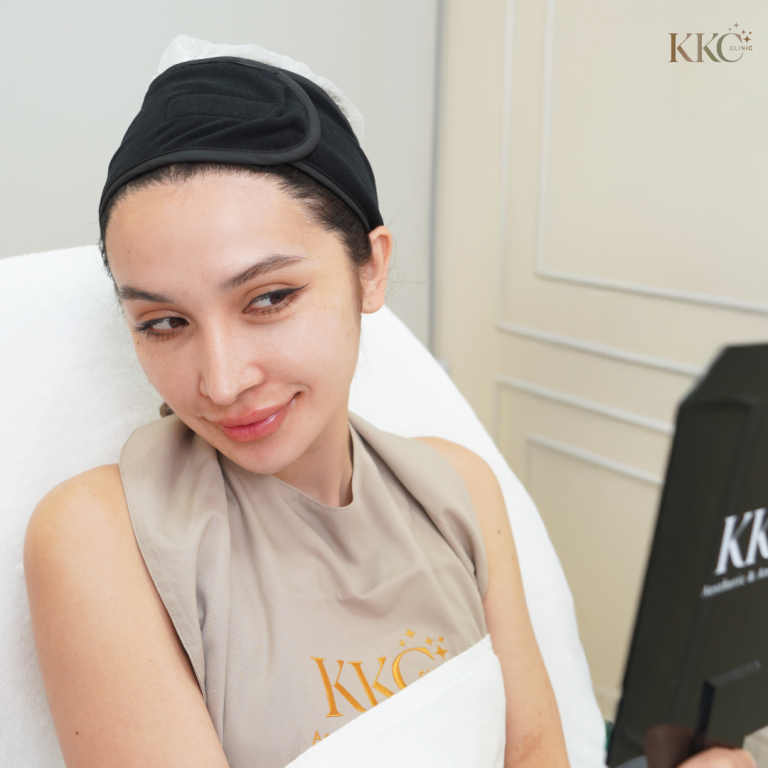หญิงสาวนั่งอยู่บนเก้าอี้ทำหัตถการ สวมผ้าคลุมของ KKC Clinic สีเบจและที่คาดผมสีดำ เธอยิ้มเล็กน้อยและมองกระจก KKC Clinic สีดำหลังจากทำหัตถการเสริมความงาม