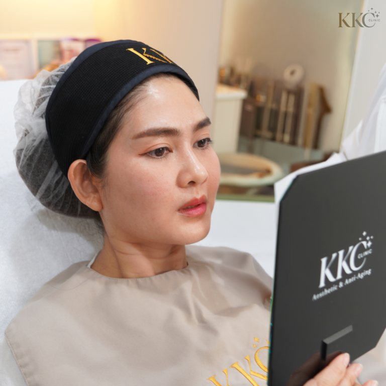 ผู้หญิงนอนบนเก้าอี้ทำทรีตเมนต์ สวมที่คาดผมและผ้าคลุมของ KKC กำลังส่องกระจกตรวจดูใบหน้าของตัวเอง