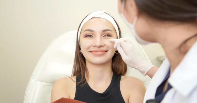 Woman smiling while a doctor in gloves examines the eye area before anti-wrinkle injections.